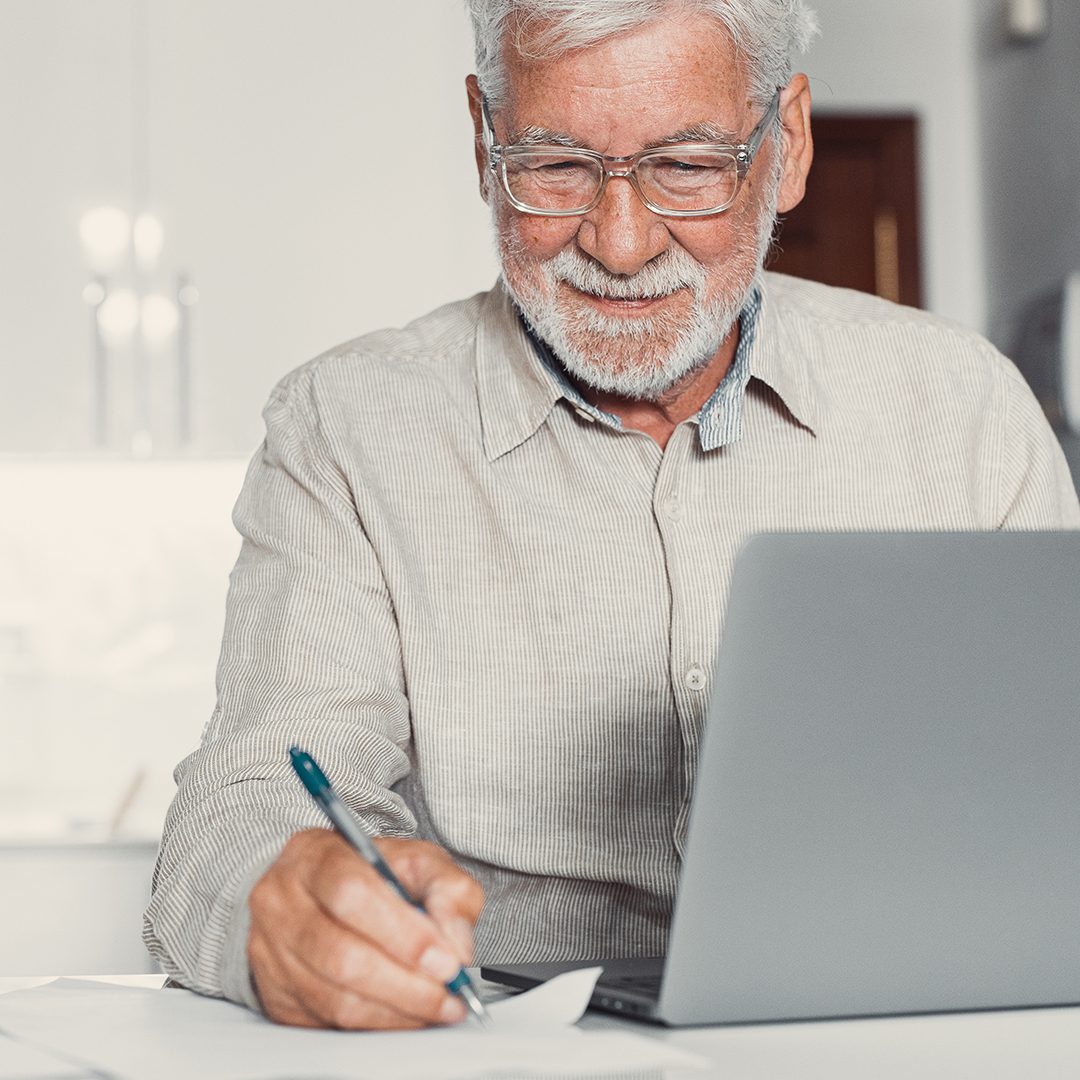An elderly internet user, using a laptop and making notes with paper and pen