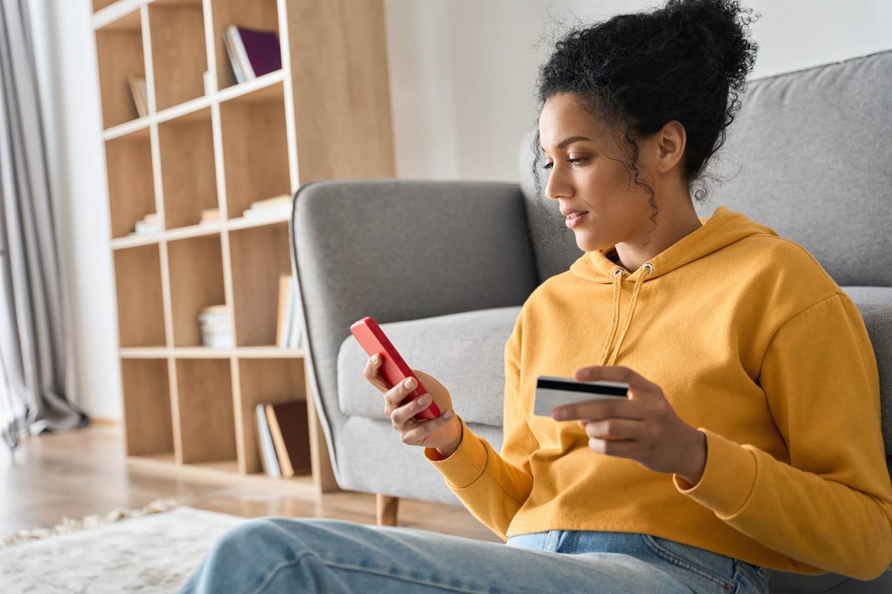 Young woman holding credit card and smartphone sitting on floor at home doing online banking transaction