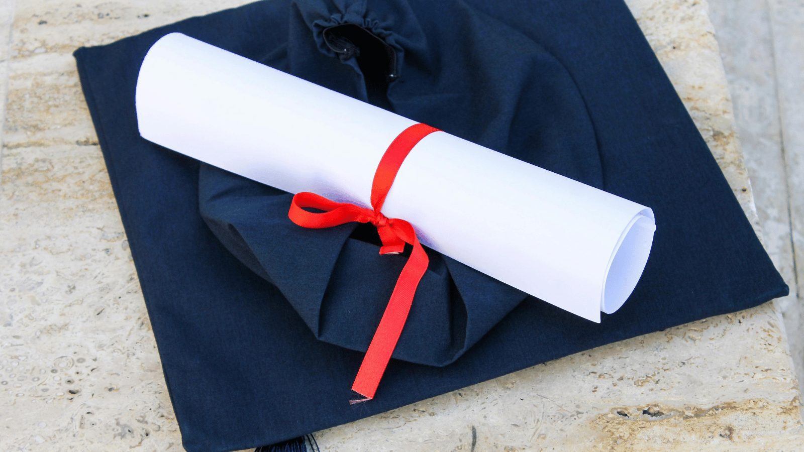 A graduation ceremony set featuring a navy blue academic cap (mortarboard) resting on a stone surface. A rolled white diploma tied with a bright red ribbon is placed diagonally across the top of the cap.