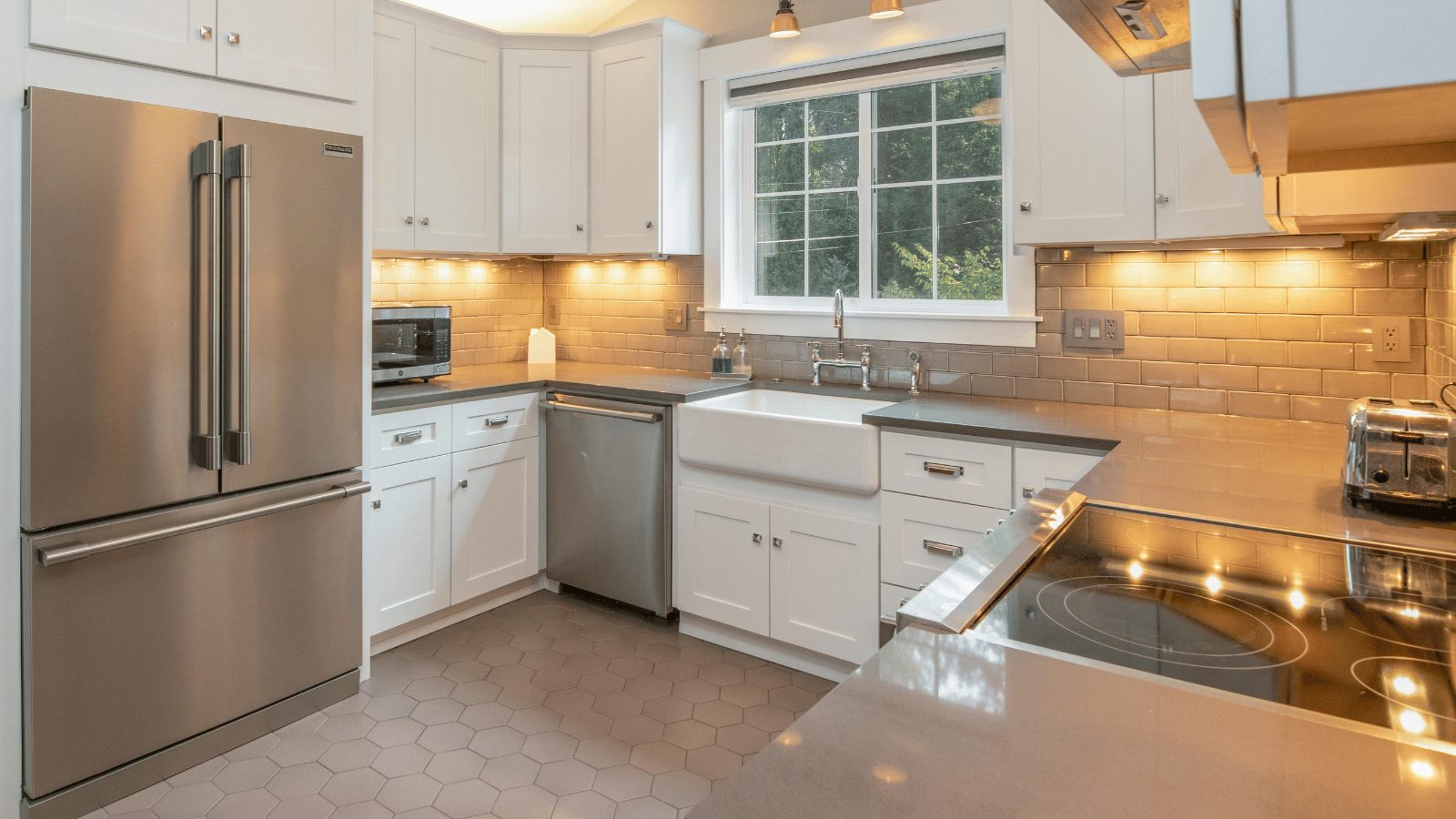 A modern, clean kitchen featuring white cabinetry, light gray countertops, and stainless steel appliances. The setup includes a large French-door refrigerator, a farmhouse-style sink under a window, and a built-in stovetop in the foreground.