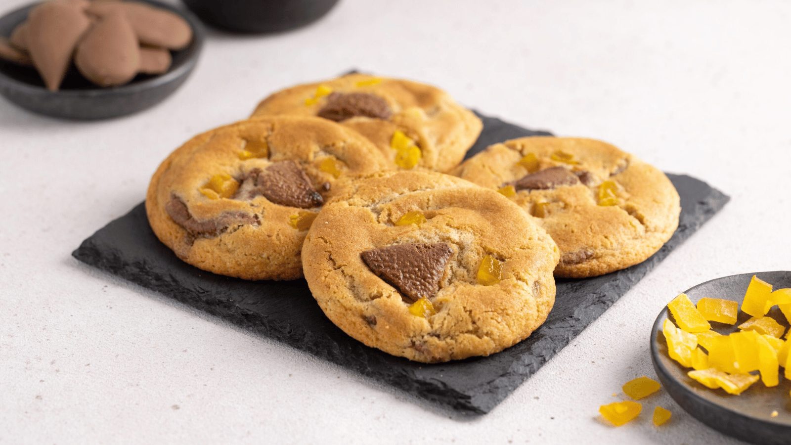 Four large chocolate chip cookies with yellow candy or fruit pieces scattered on top. They are served on a dark slate tray, accompanied by small bowls of extra chocolate and yellow toppings.