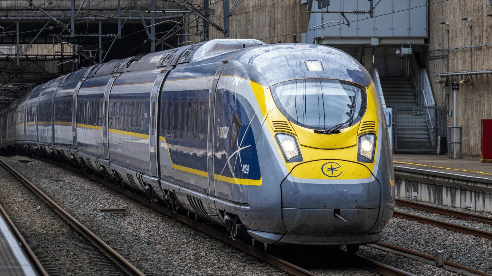 A sleek, modern high-speed Eurostar train stopped at a station platform. The train is gray with a bright yellow front and blue accents along the sides.