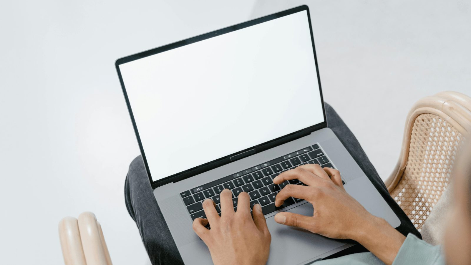 An over-the-shoulder view of a person using a laptop while sitting in a wicker chair. The laptop screen is blank white, and the person's hands are positioned over the keyboard.
