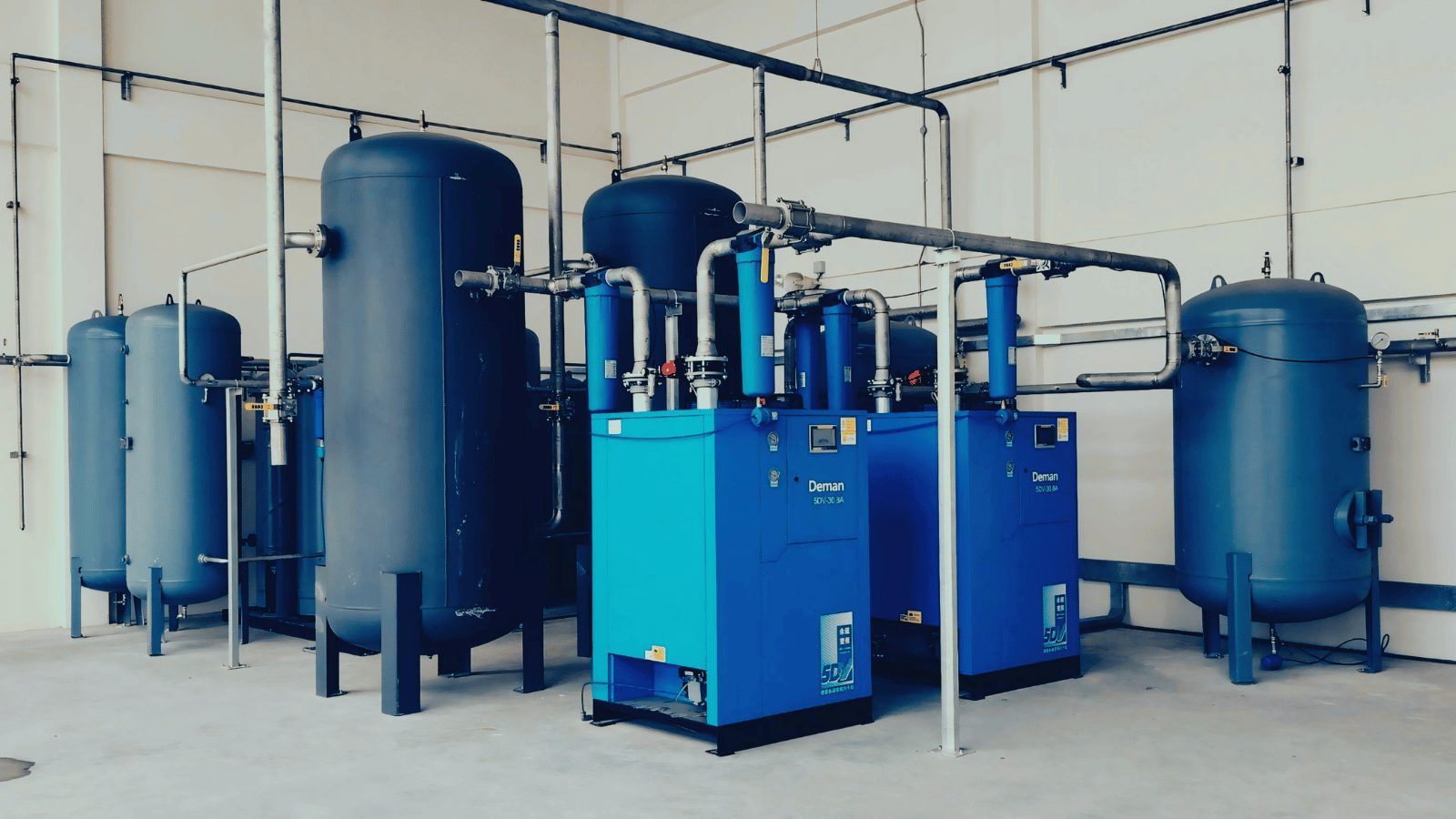 An industrial utility room filled with large blue machinery and storage tanks. Complex silver piping connects the various units, which appear to be part of a compressed air or water filtration system.