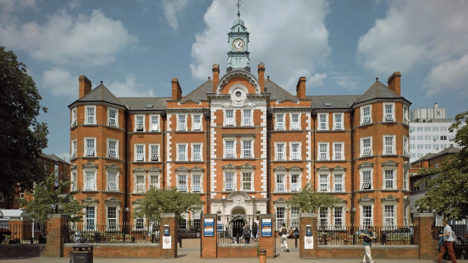 A large, historic red-brick building with multiple windows and a central clock tower. The architecture is grand and symmetrical, featuring white stone trim around the windows and entrance.