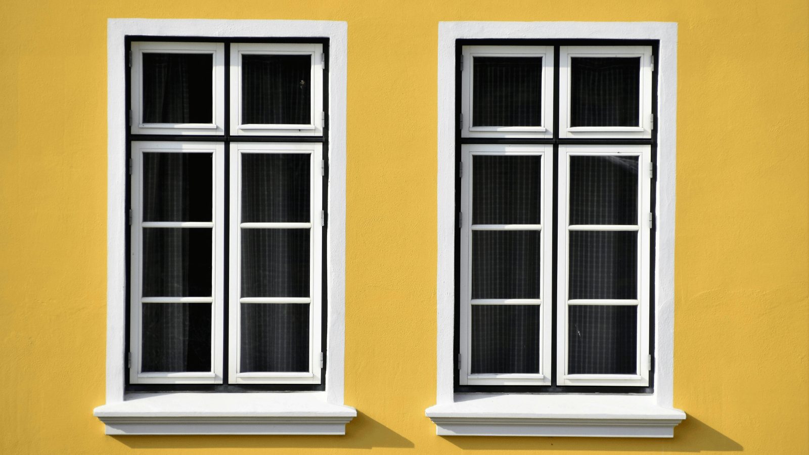 A close-up of two identical rectangular windows set into a bright yellow wall. The windows have white frames and black grilles, with dark curtains visible inside.