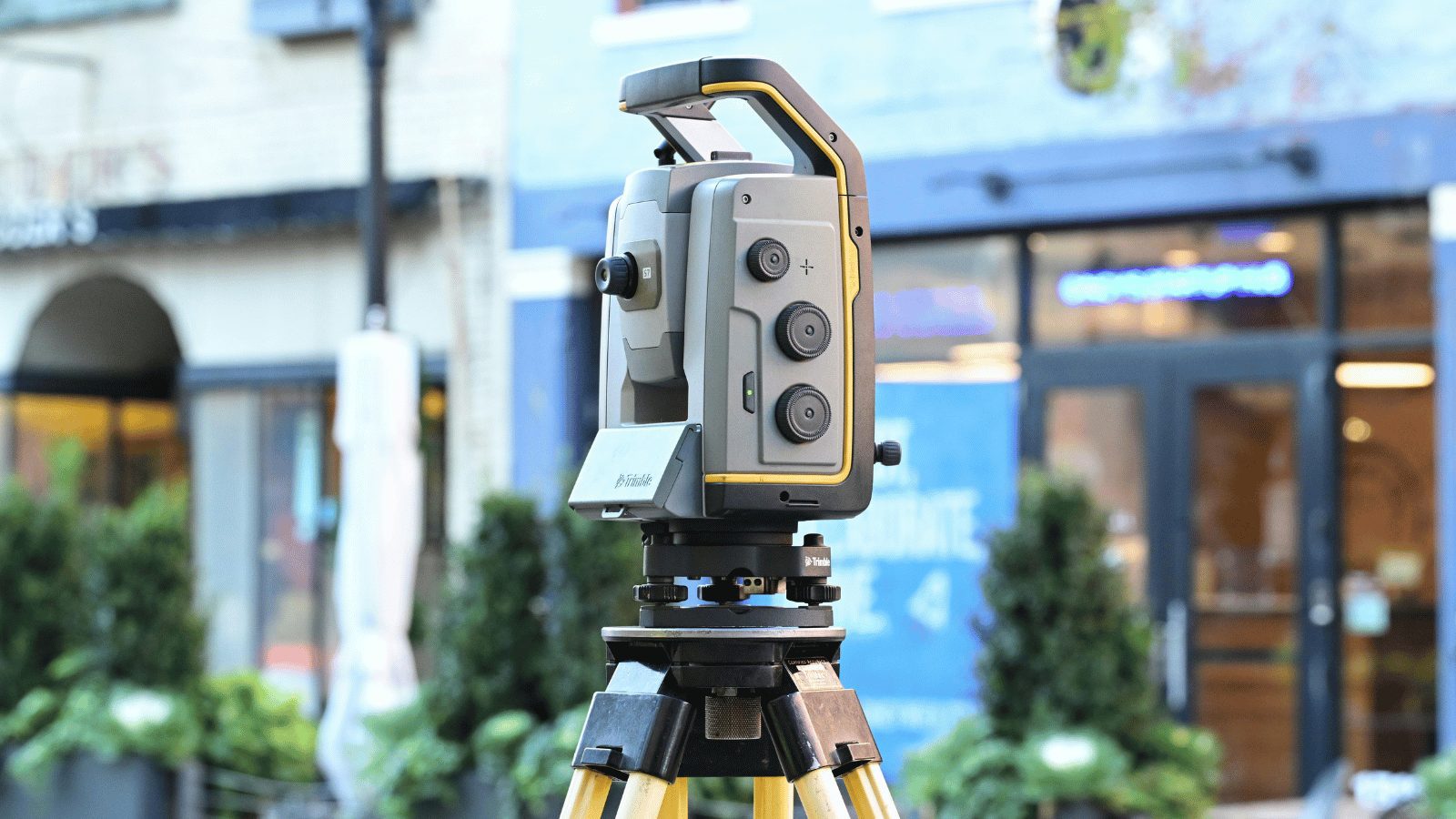 A professional surveying instrument (total station) mounted on a yellow tripod in an urban outdoor setting. The background shows blurred storefronts and greenery.