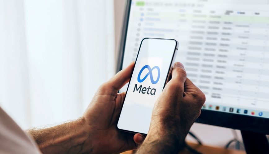 Hands holding photo with Meta logo on screen. Computer screen in the background with Blurred spreadsheet open