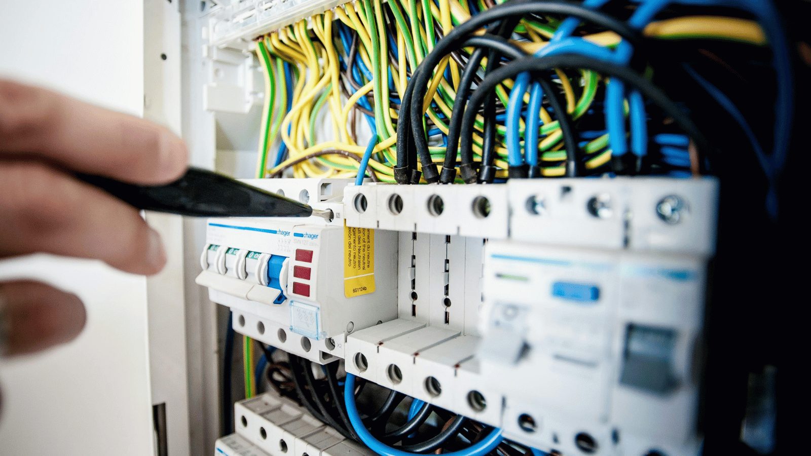 A close-up of a person’s hand using a black tool to work on a complex electrical circuit breaker panel. The panel is filled with numerous organized wires in green, yellow, and blue.