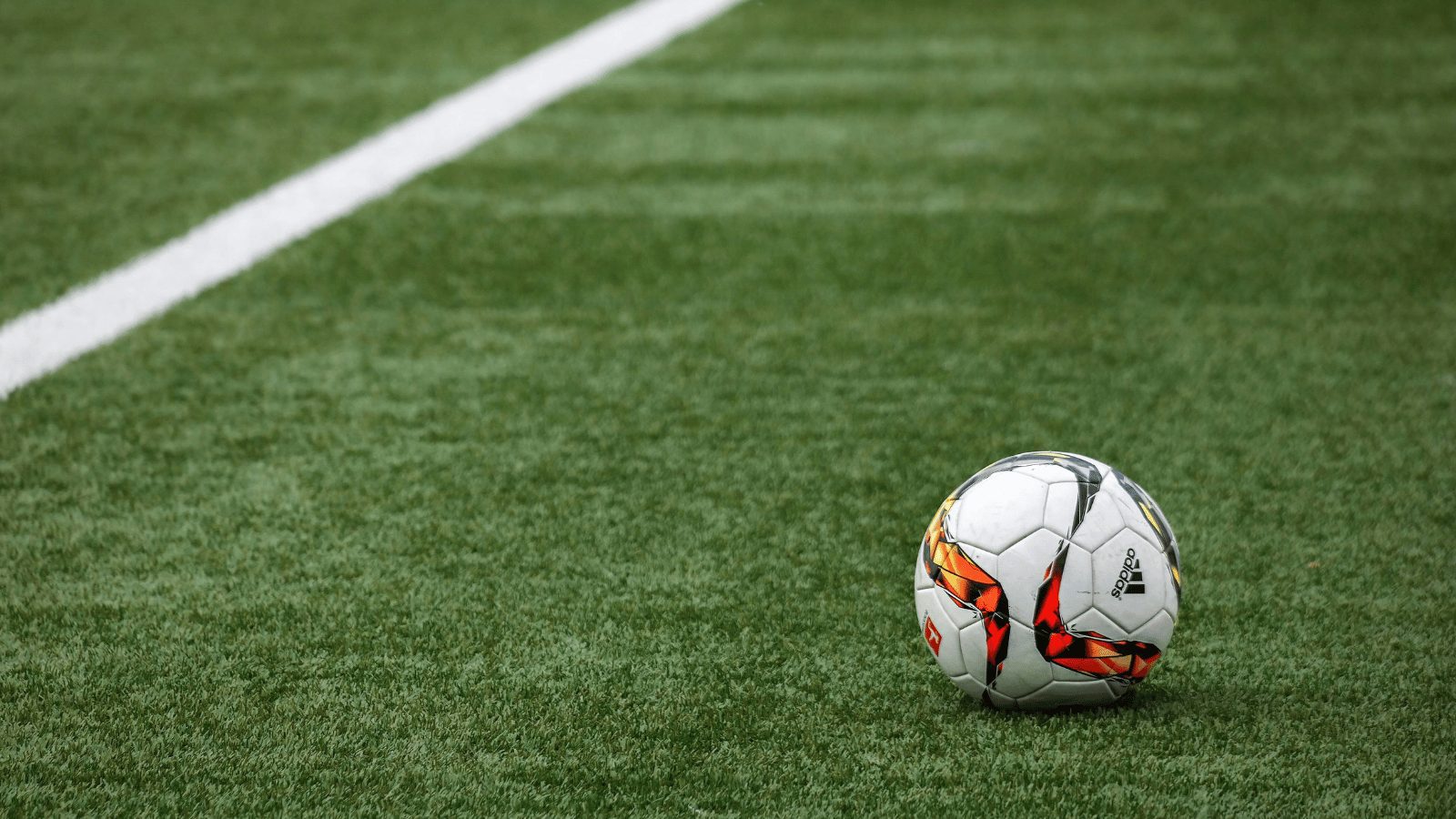 A close-up of a white soccer ball resting on green grass. The ball features a modern design with bold orange and black curved graphic patterns.