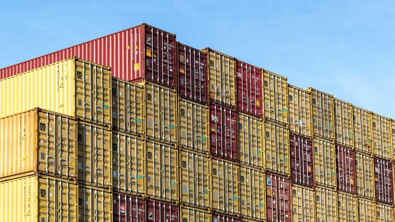 A massive stack of multicolored shipping containers piled high against a clear blue sky. The containers are mostly yellow and deep red/burgundy, arranged in a dense, industrial grid.
