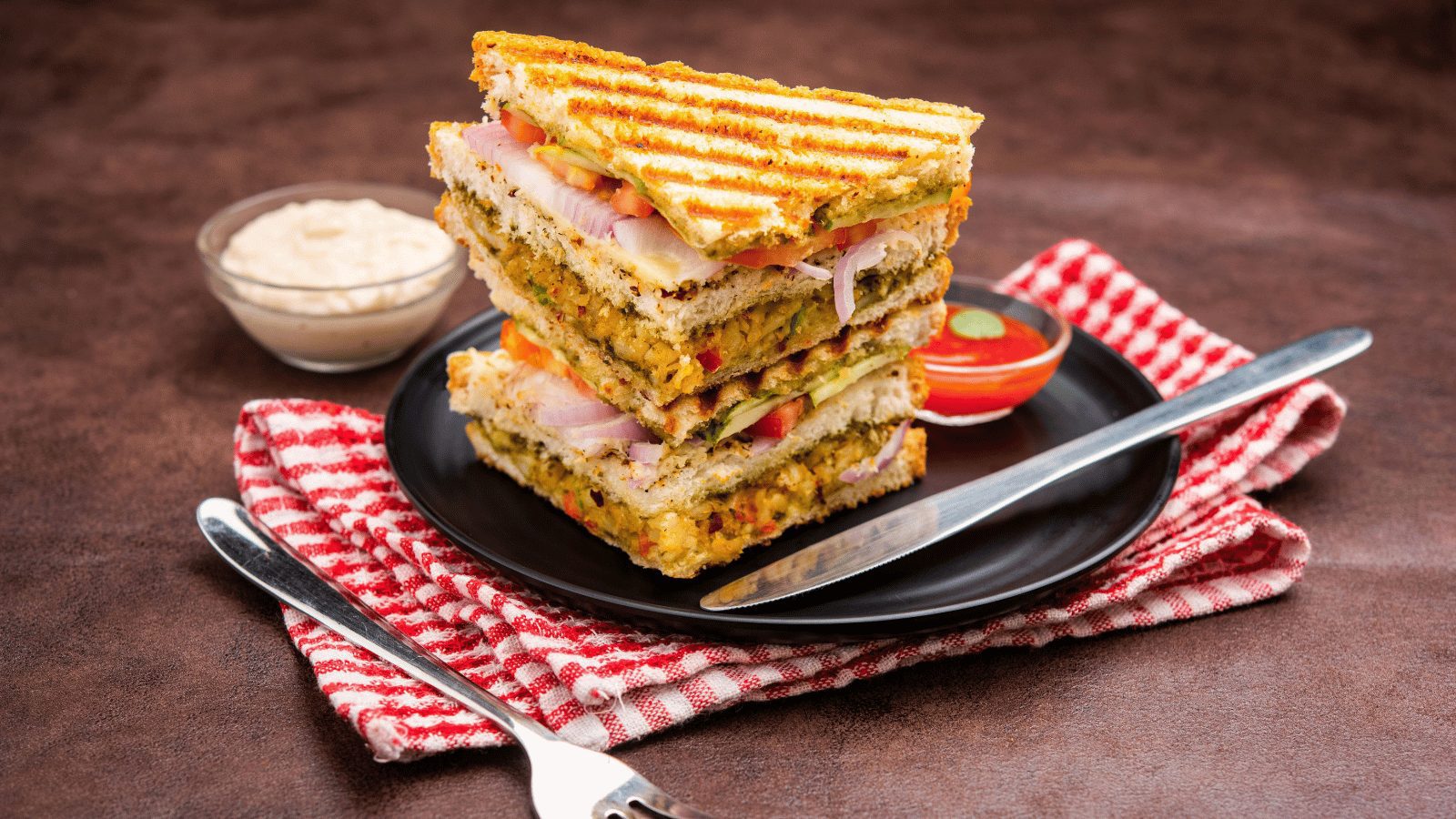A tall, multi-layered club sandwich cut into triangles and stacked on a black plate. The sandwich is toasted and filled with vegetables and sauces, served with small side dishes of dip.
