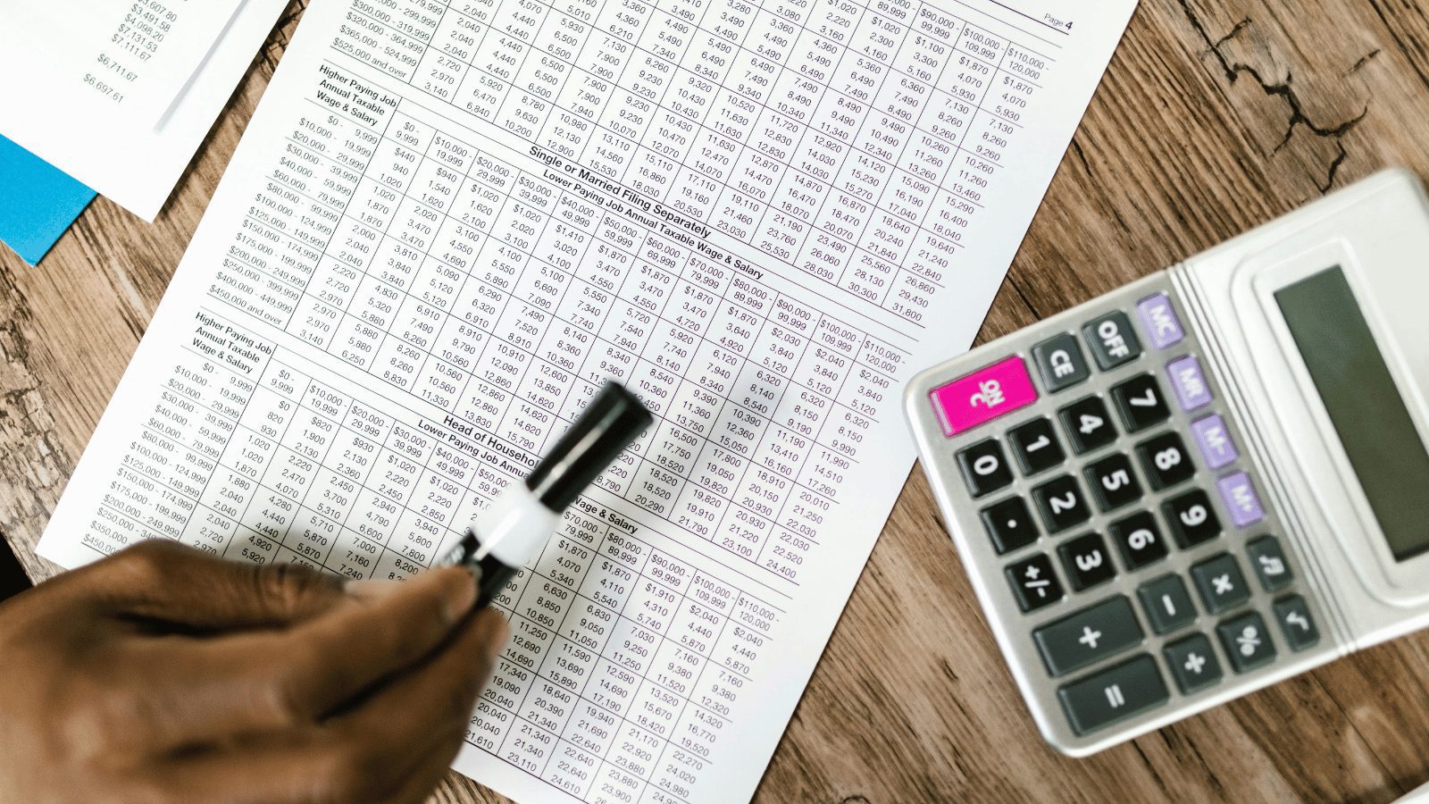 Zoomed in on table with spreadsheets with lots of numbers on it and a calculator on it. A person pointing at the sheet with Black pen.