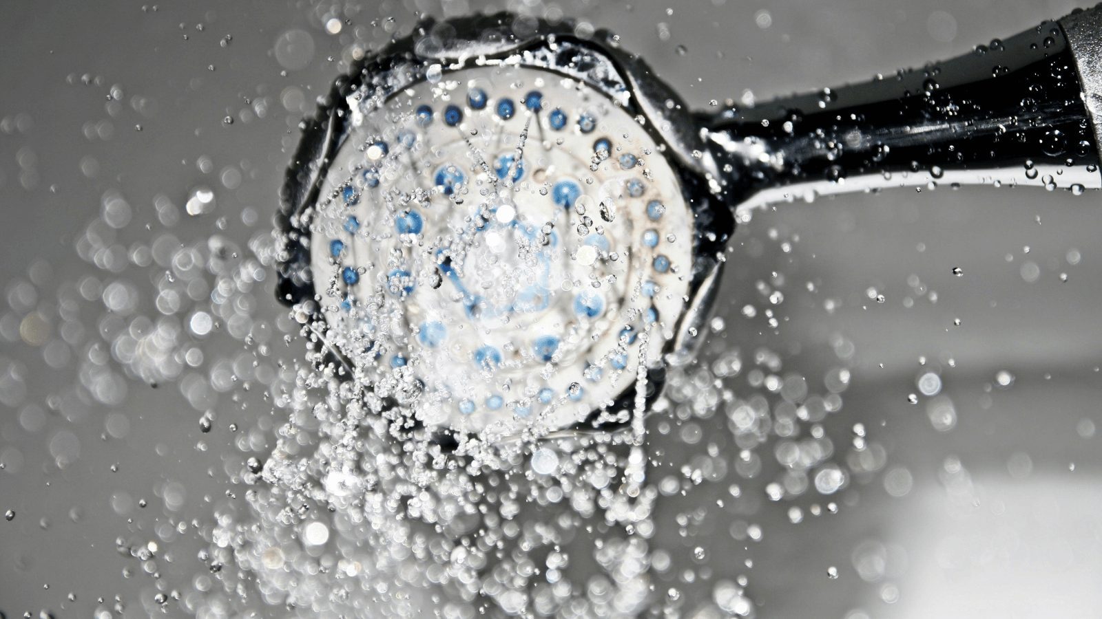 A high-speed action shot of a chrome shower head with water spraying out in many directions. The background is blurred, focusing on the droplets and the flow of the water.